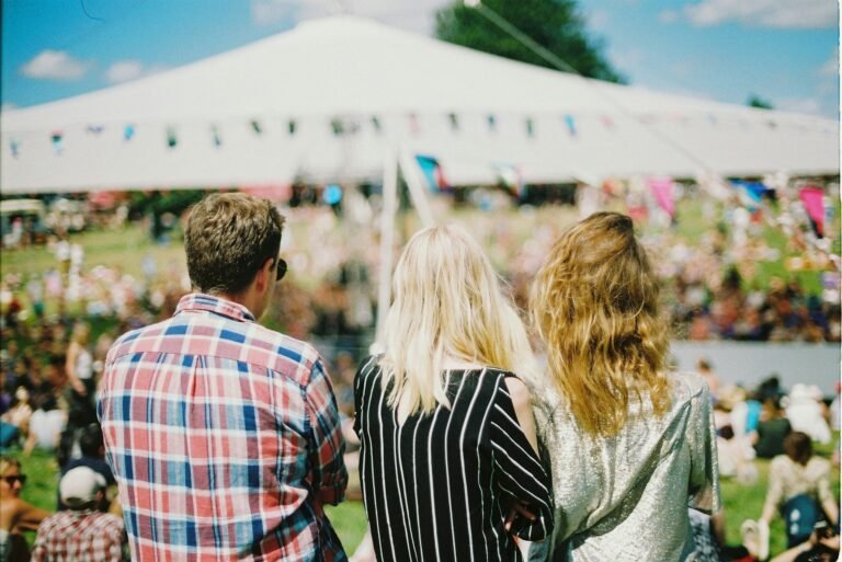 Three people seen from the back, standing in a field on a sunny day, looking towards a large tent with a crowd of people gathered at an outdoor event.